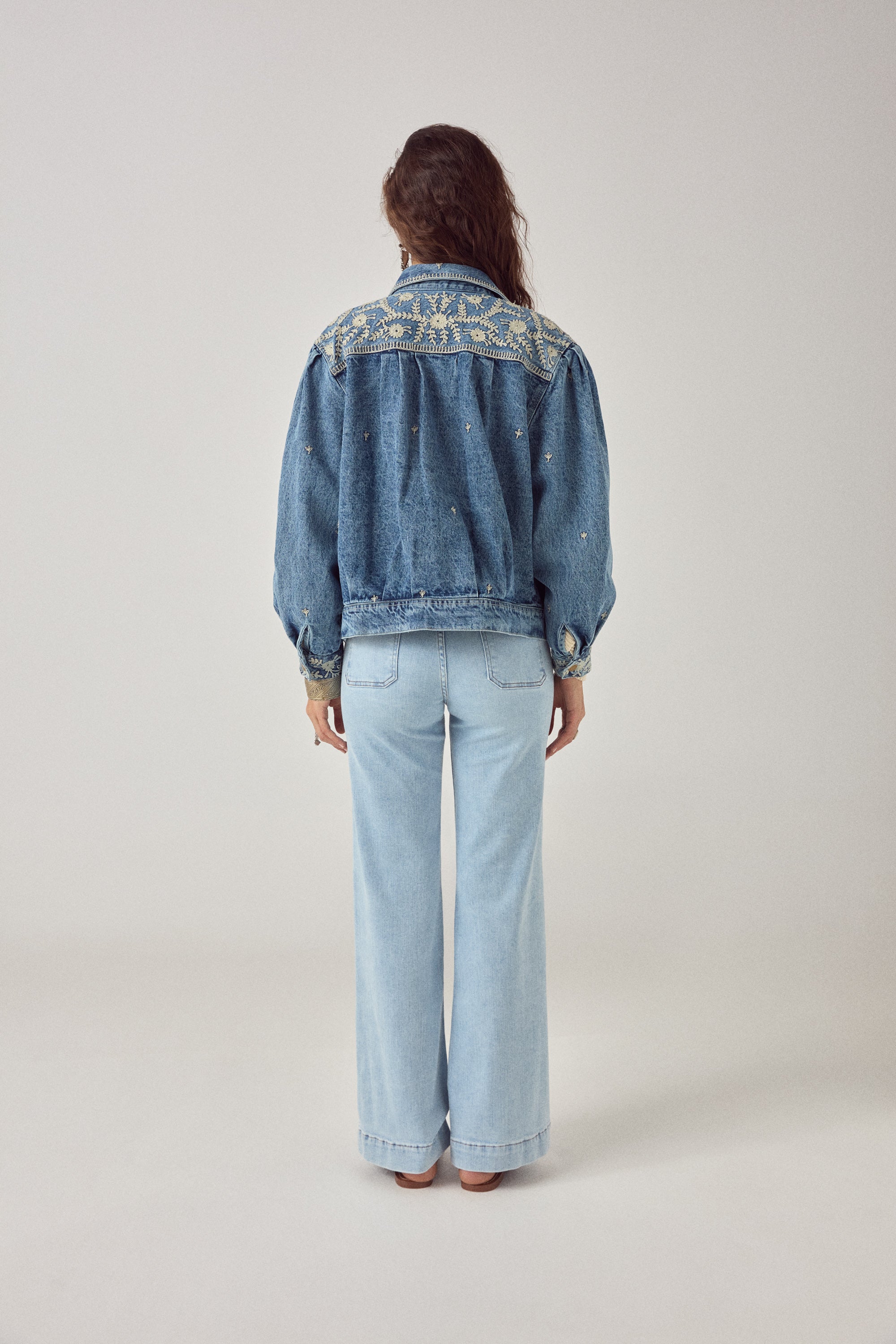 Woman wearing a blue denim jacket with white floral embroidery detailing with denim jeans on a plain background