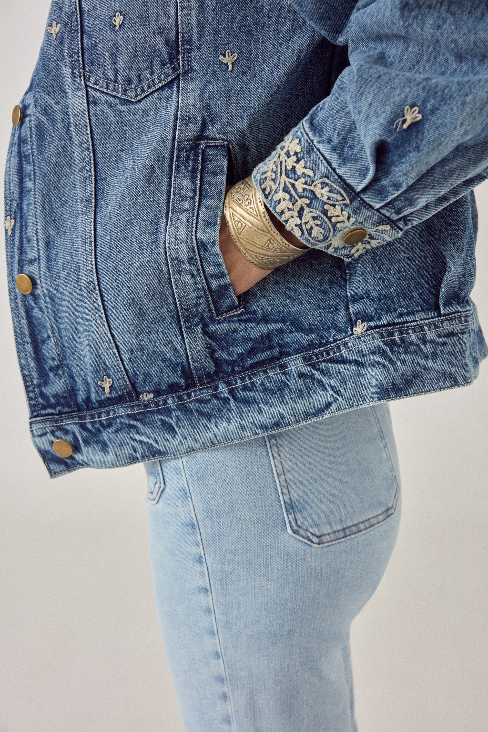 Woman wearing a blue denim jacket with white floral embroidery detailing with denim jeans on a plain background