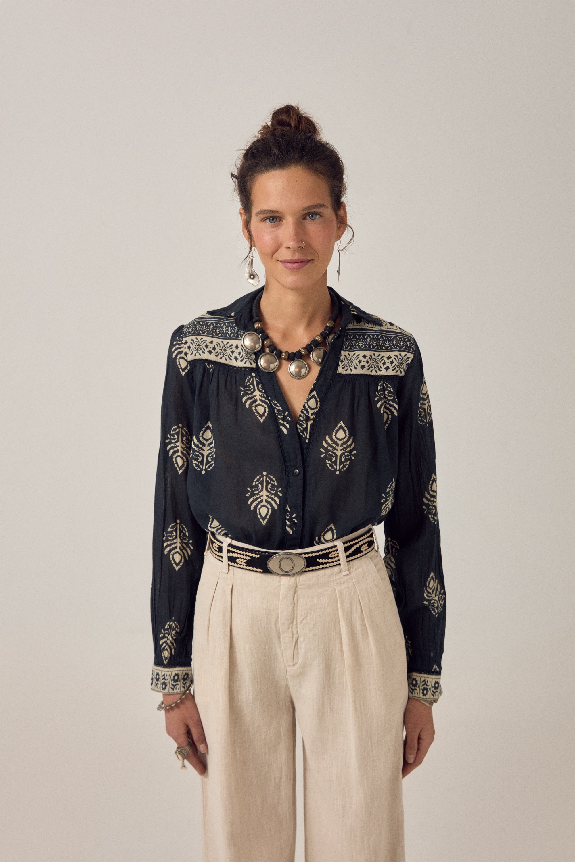 Woman wearing a black embroidered blouse with beige pants on a plain background