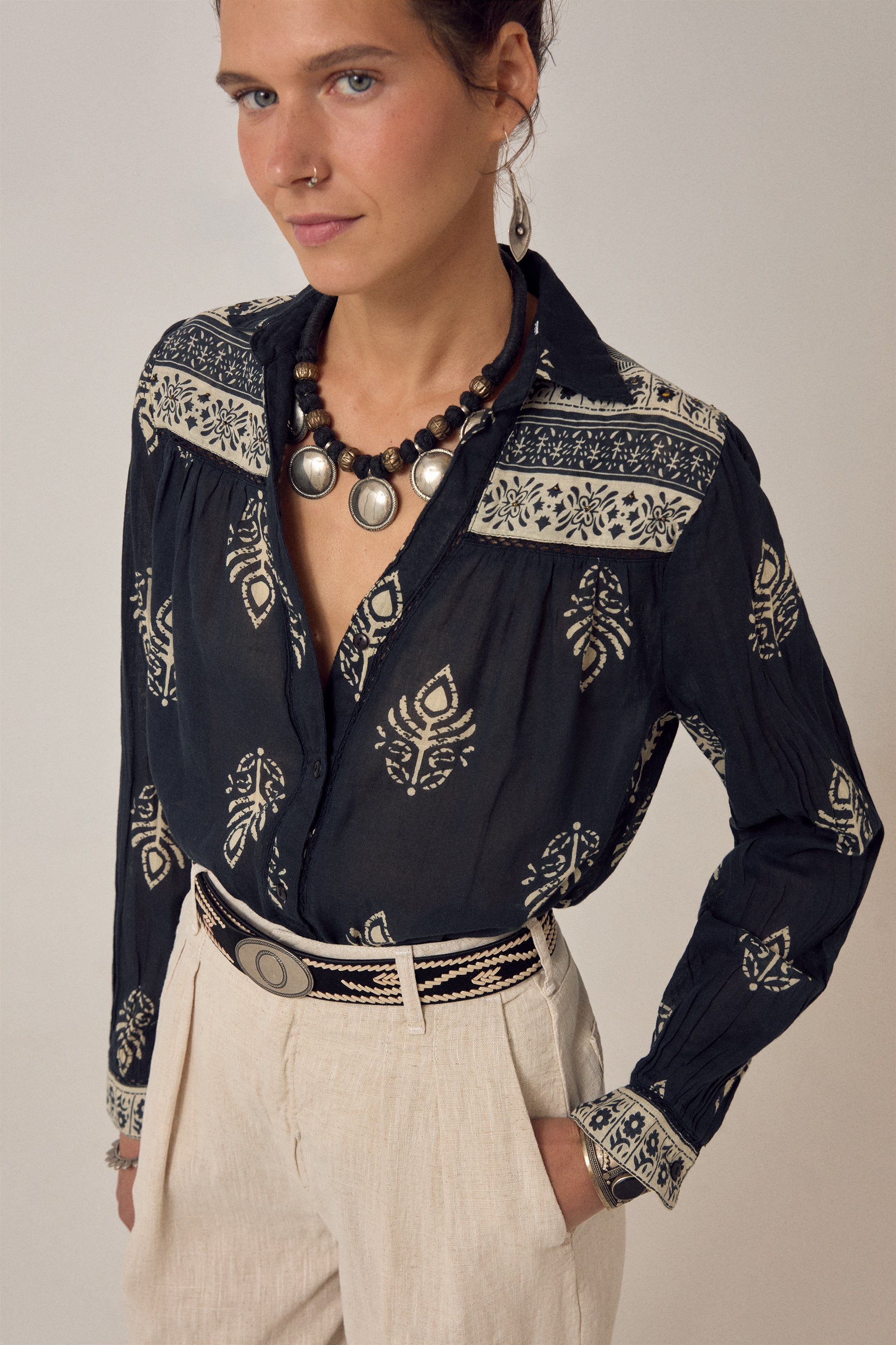 Woman wearing a black embroidered blouse with beige pants on a plain background