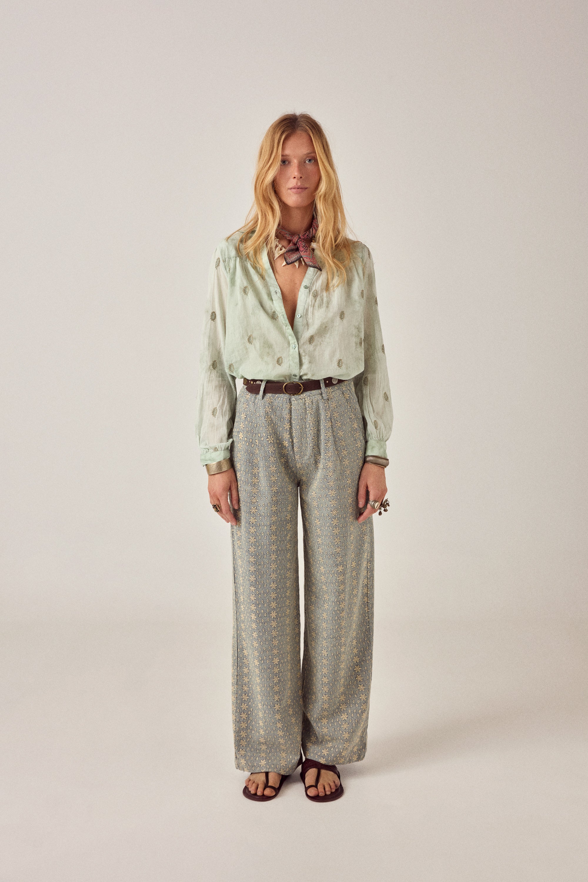 Woman wearing a light green blouse with floral patterns and patterned pants on a plain background