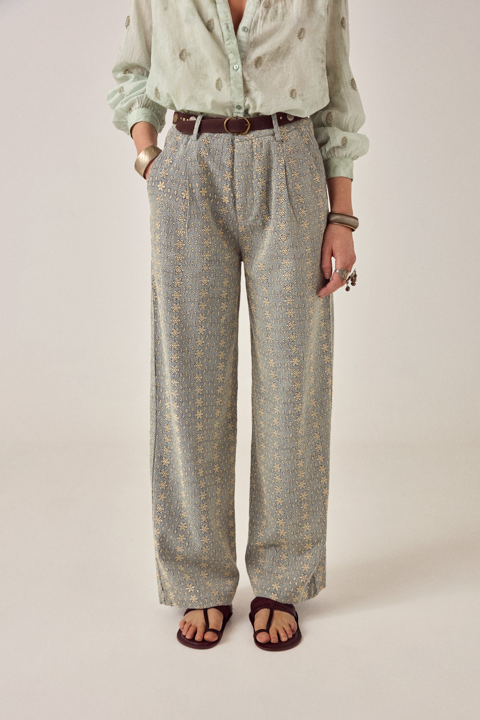 Woman wearing a light green blouse with floral patterns and patterned pants on a plain background