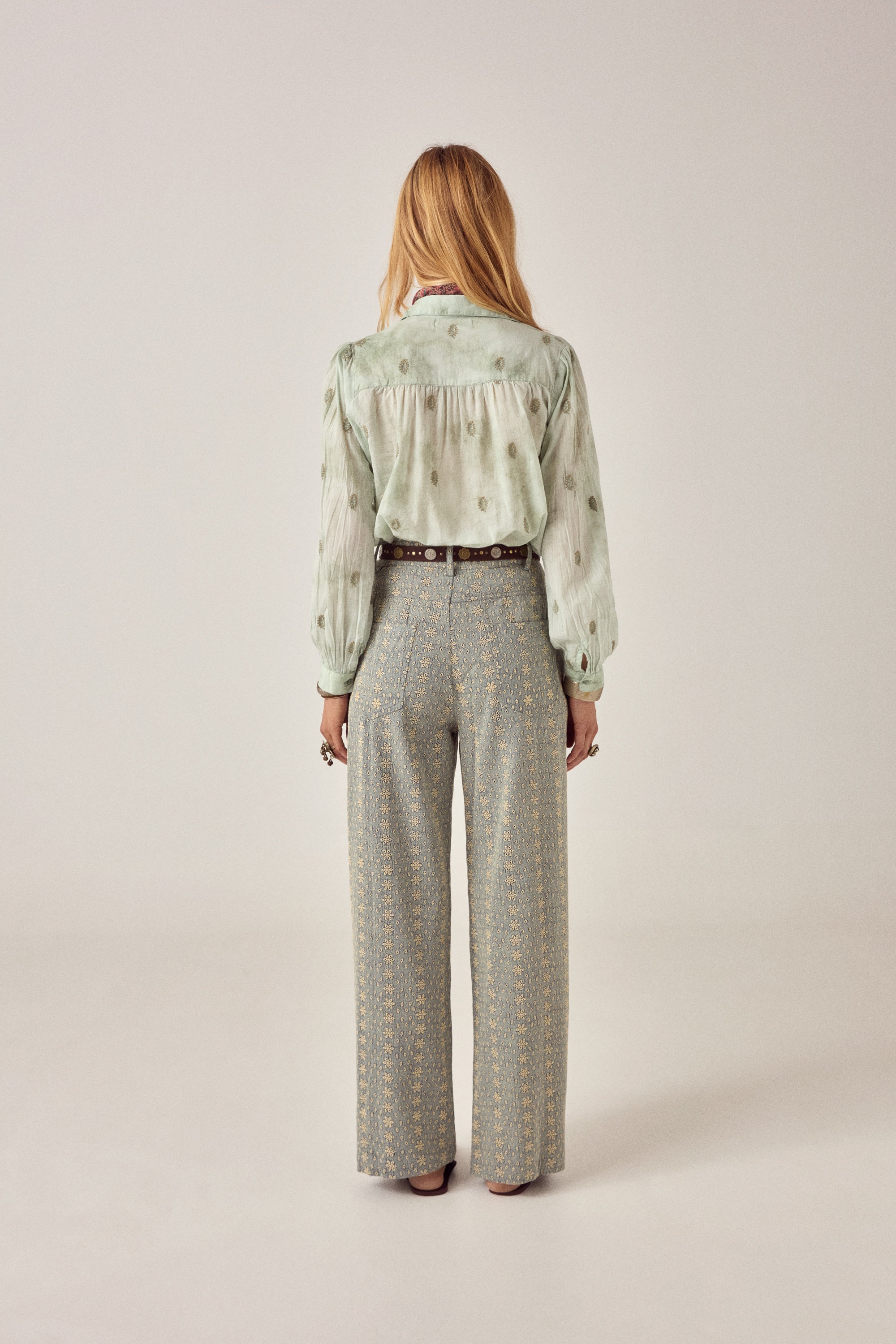Woman wearing a light green blouse with floral patterns and patterned pants on a plain background