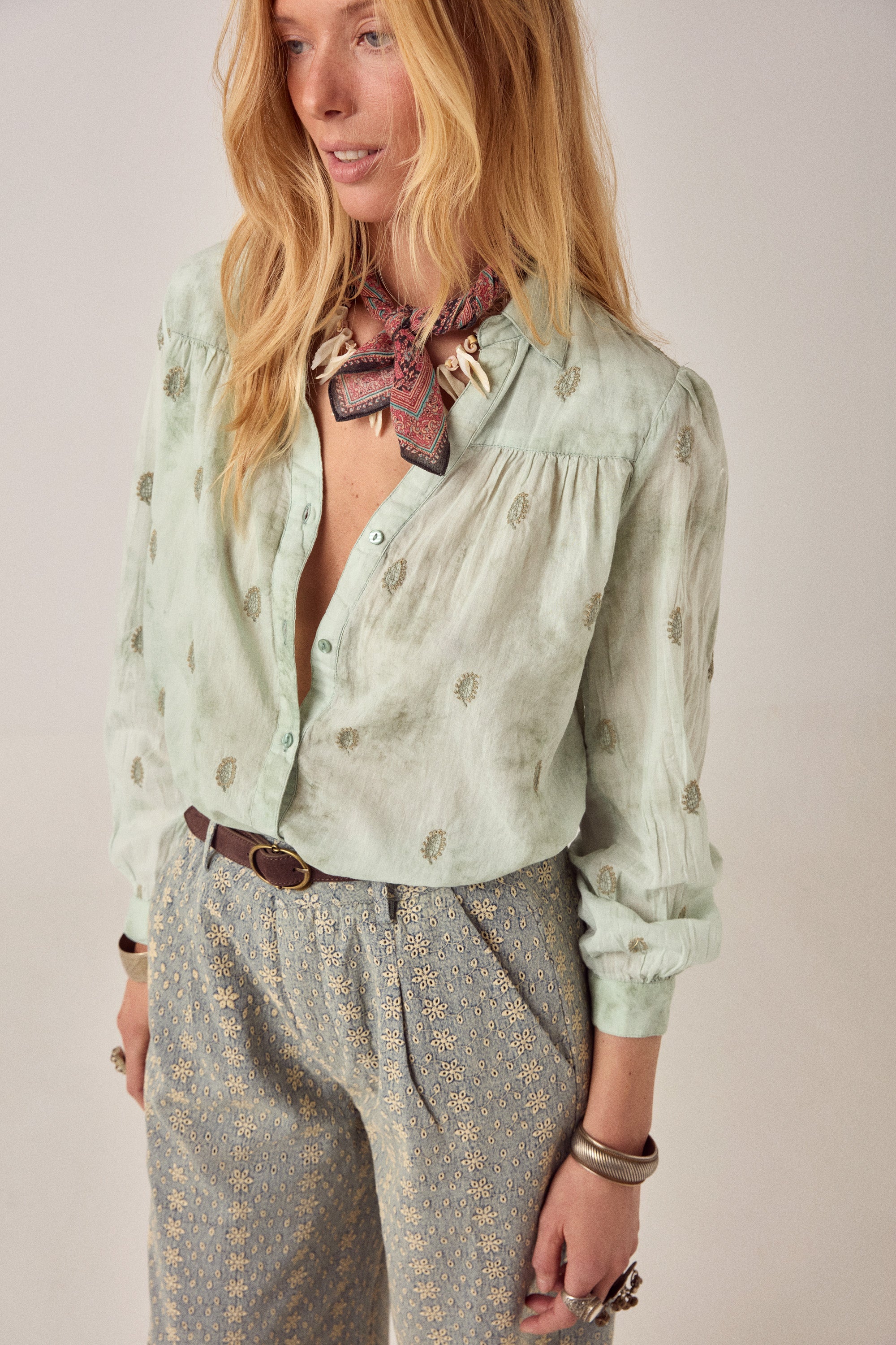 Woman wearing a light green blouse with floral patterns and patterned pants on a plain background