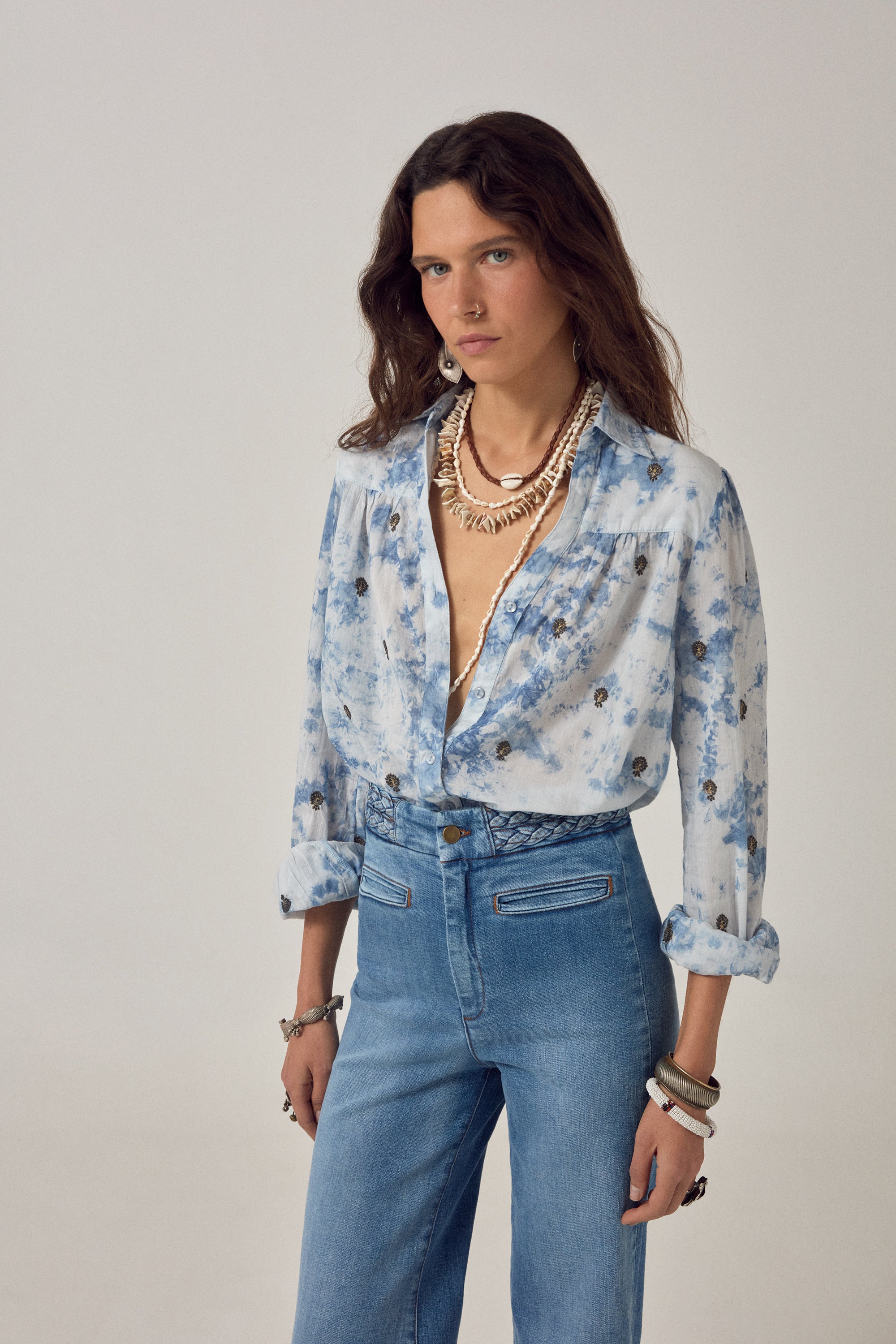 Woman wearing a blue and white blouse with embroidered detail and denim jeans on a white background. 