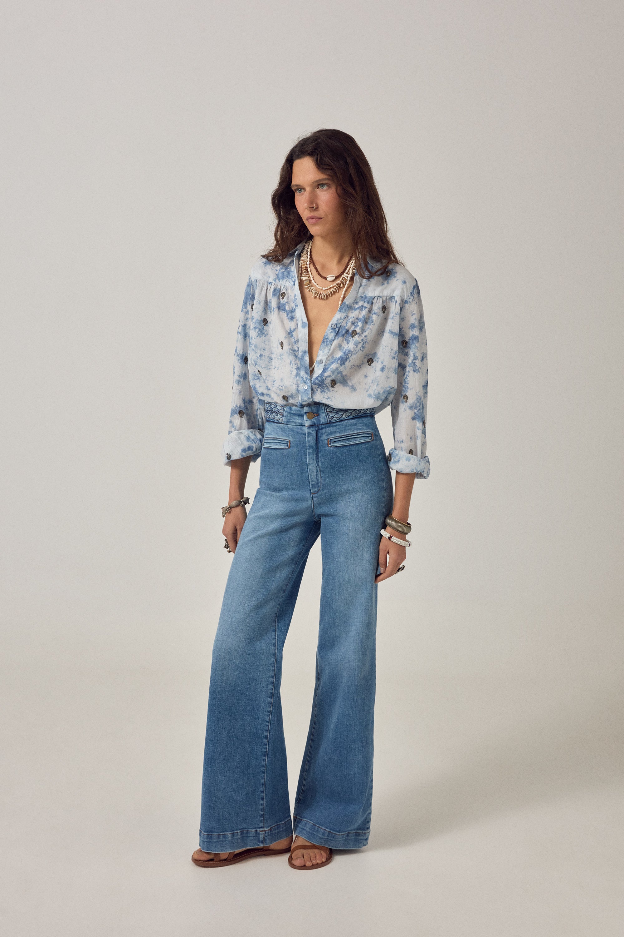 Woman wearing a blue and white blouse with embroidered detail and denim jeans on a white background. 