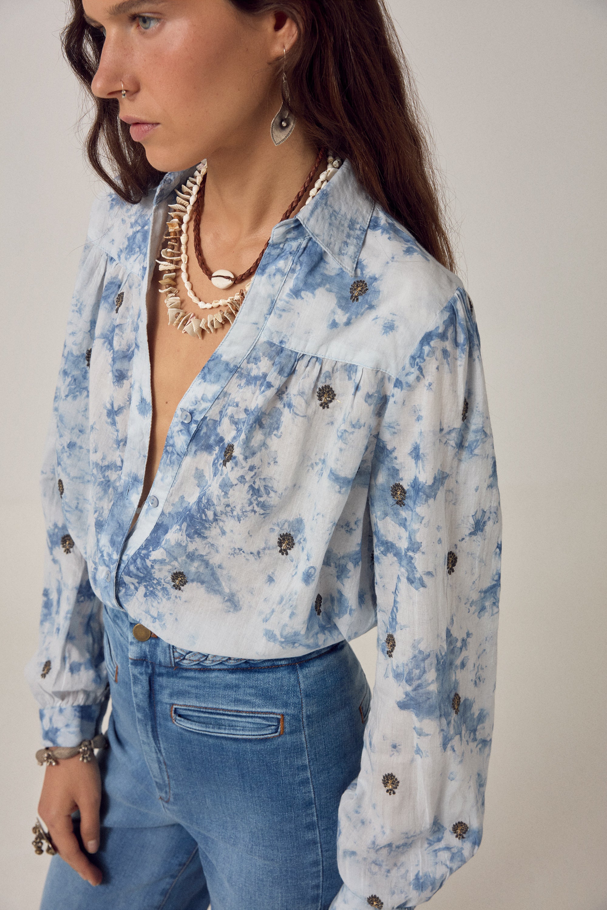 Woman wearing a blue and white blouse with embroidered detail and denim jeans on a white background. 