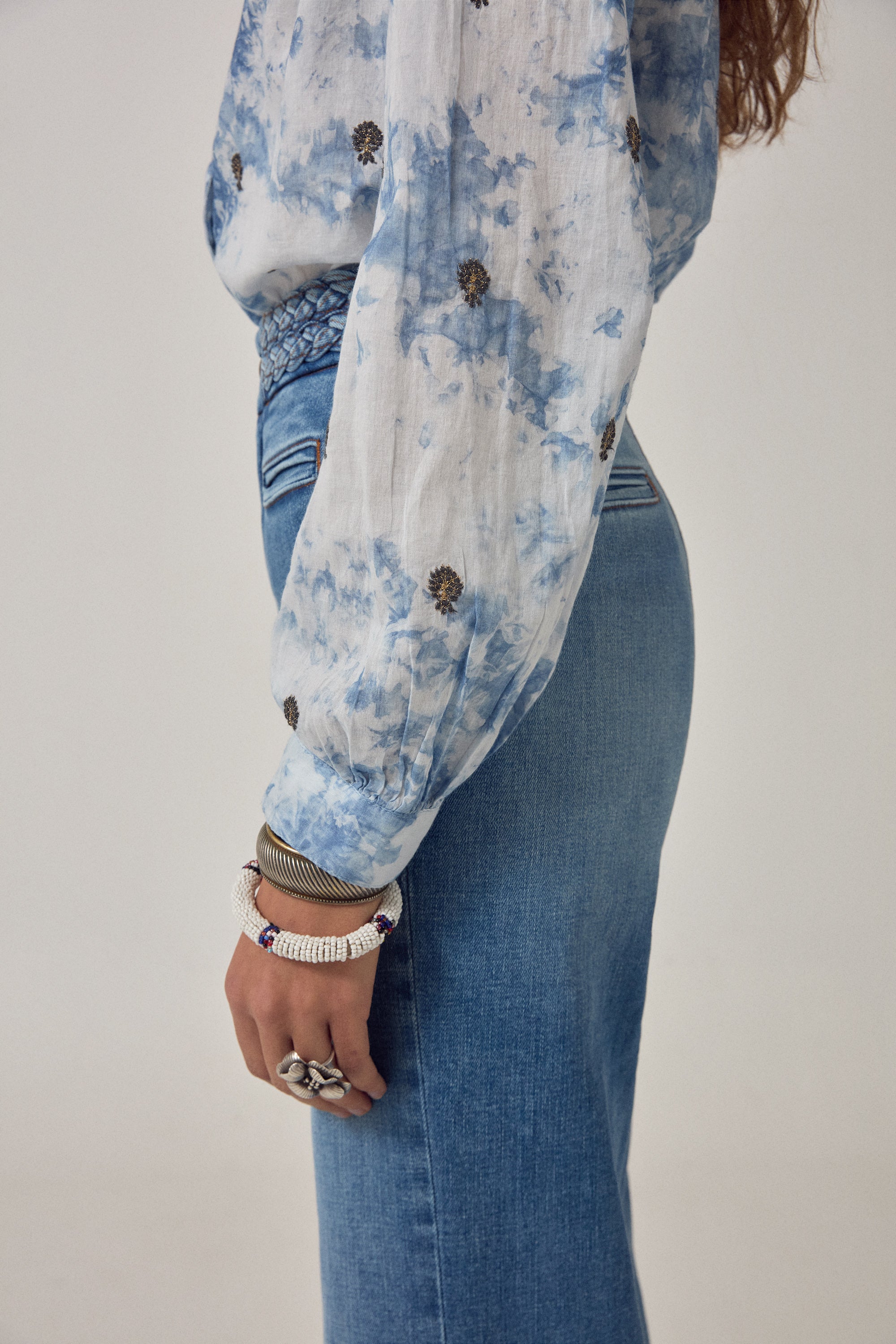 Woman wearing a blue and white blouse with embroidered detail and denim jeans on a white background. 