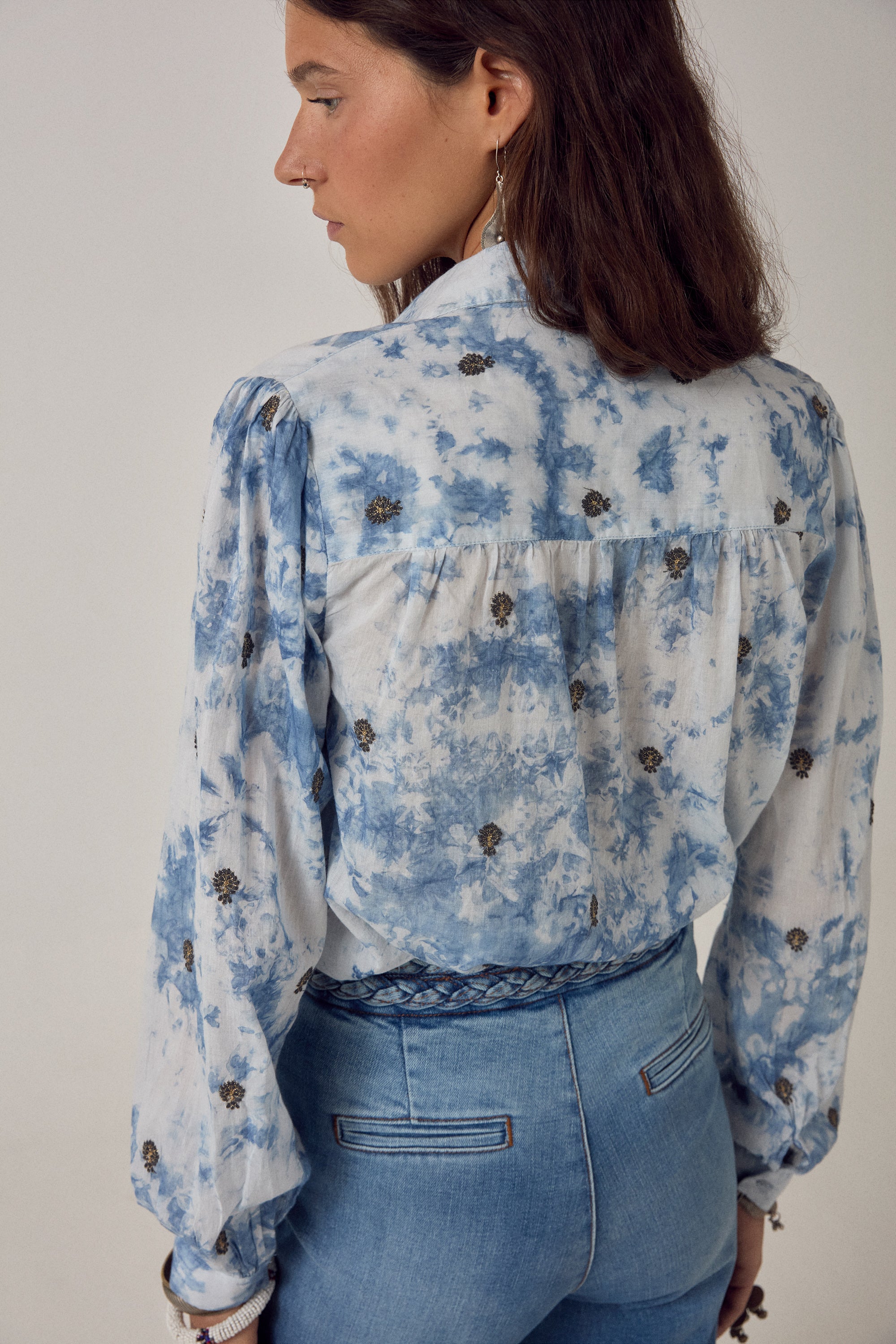 Woman wearing a blue and white blouse with embroidered detail and denim jeans on a white background. 