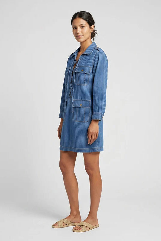 A woman modeling a blue denim shirt dress with vertical pinstripes, standing against a light gray studio background.