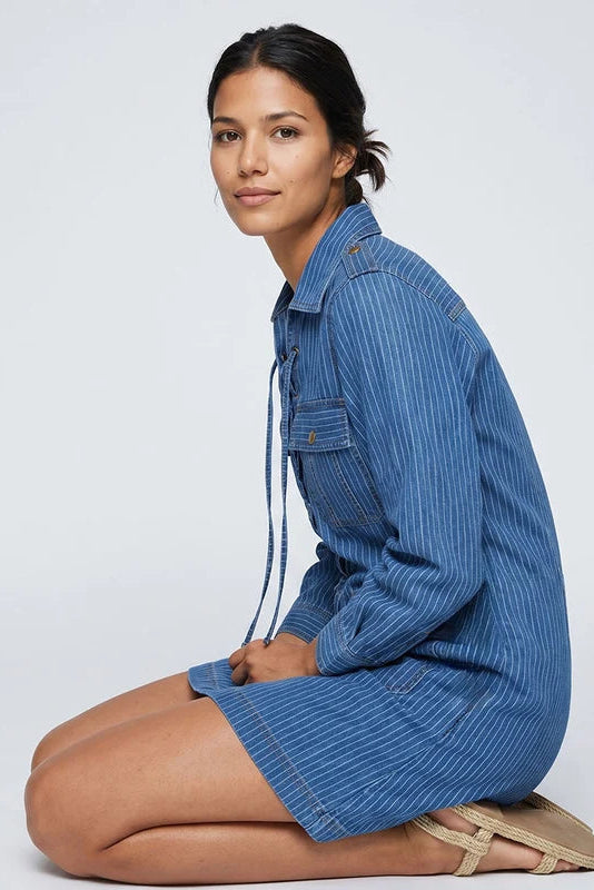 A woman kneels in a studio wearing a blue denim shirt dress with thin white vertical pinstripes. The dress has a button-down front, chest pocket, and drawstring tie.