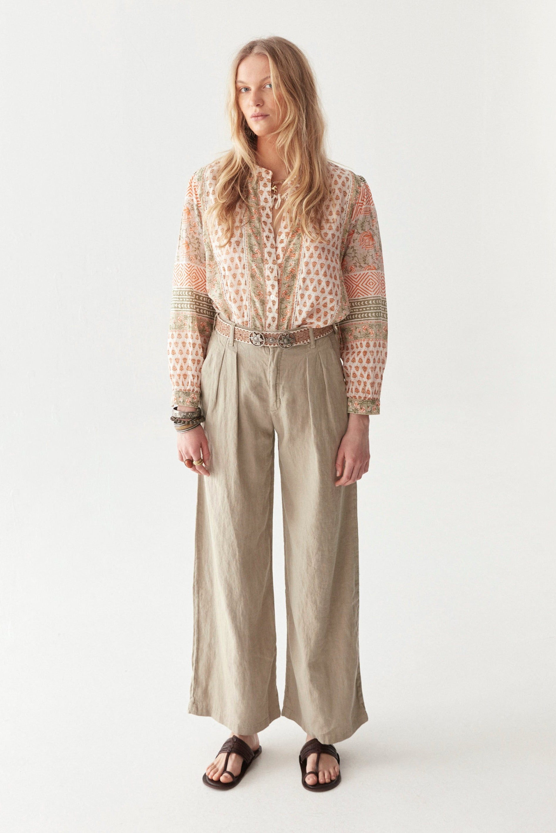 Model wearing bohemian floral shirt with long sleeves in orange and green pattern. Paired with beige wide leg pants and sandals.  

