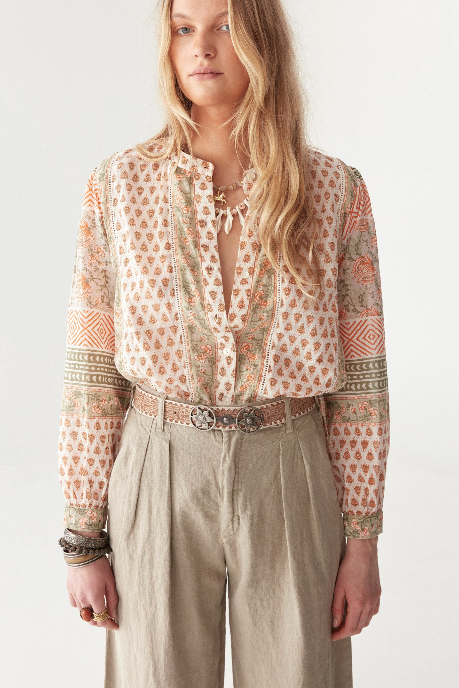 Model wearing bohemian floral shirt with long sleeves in orange and green pattern. Paired with beige wide leg pants and sandals.  
