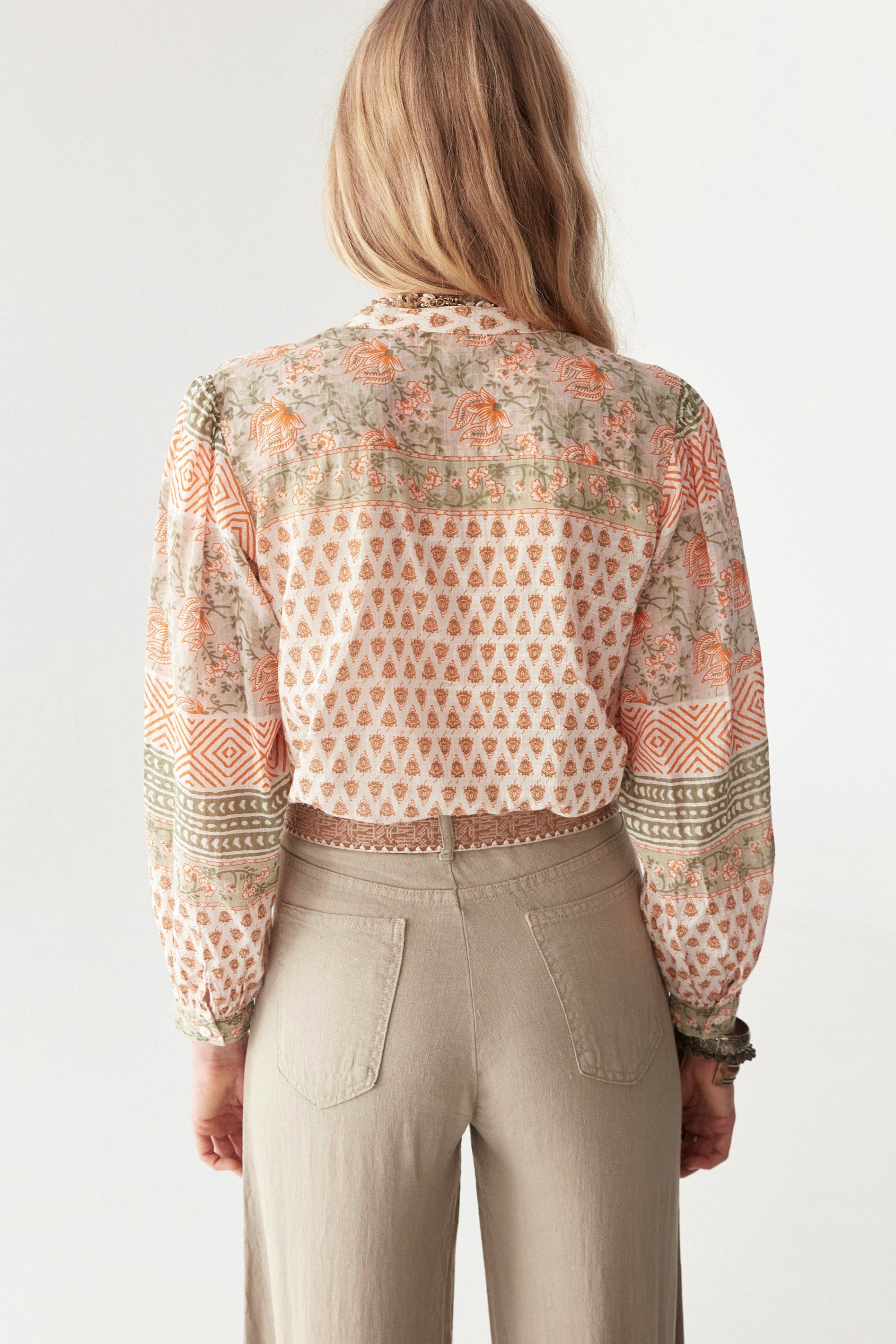 Model wearing bohemian floral shirt with long sleeves in orange and green pattern. Paired with beige wide leg pants and sandals.  
