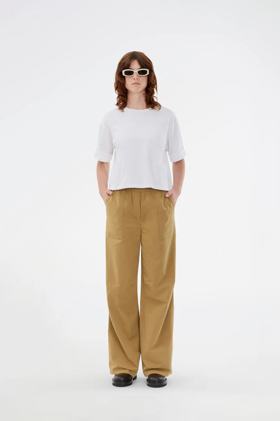 A model stands against a plain white studio background wearing a white short-sleeve crewneck t-shirt, wide-leg tan pants, and black shoes.