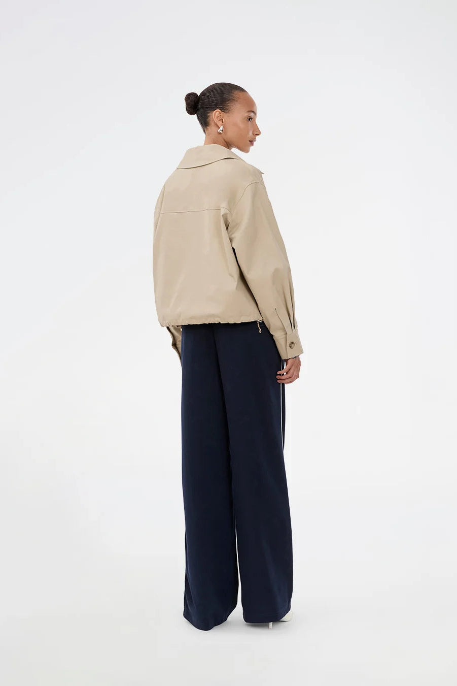 A model shown from the back wearing a beige utility-style jacket and navy wide-leg trousers, standing against a plain white studio background.