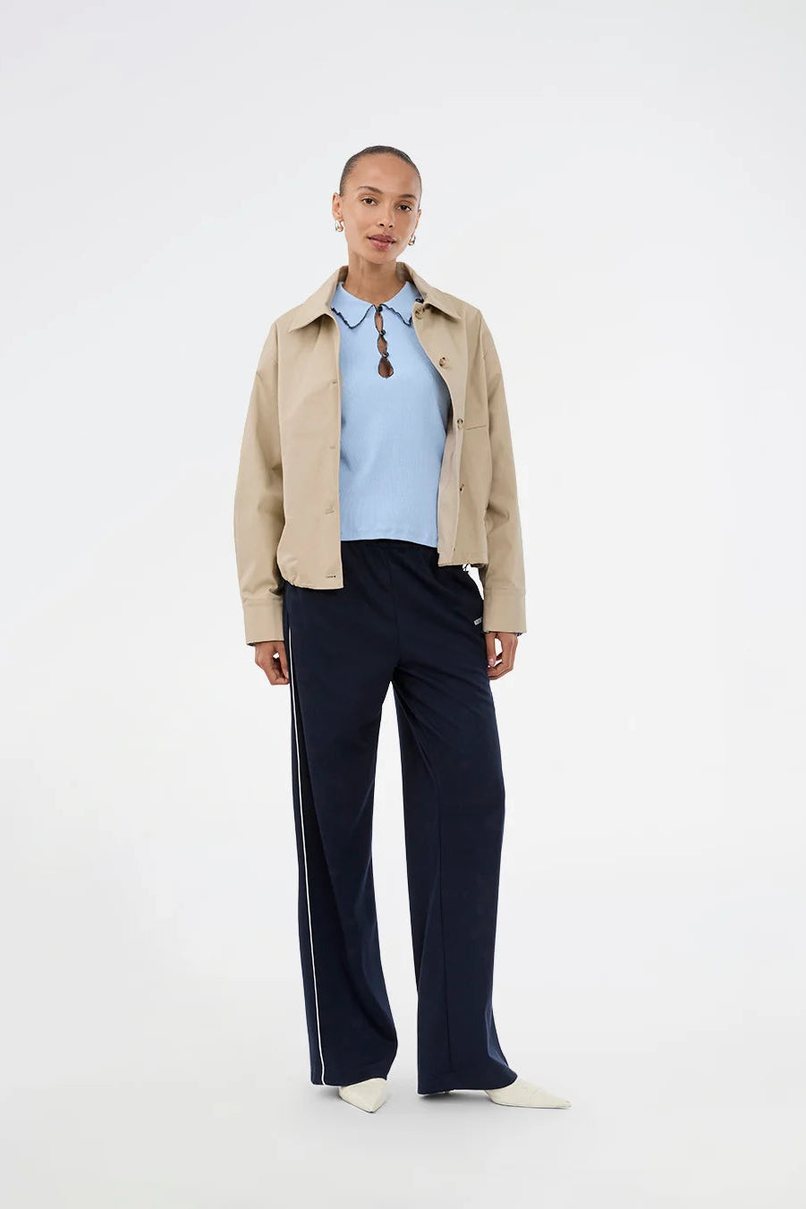 A model stands against a plain white studio background wearing a light beige cropped jacket over a light blue polo shirt, paired with dark navy wide-leg track pants featuring a white side stripe, and white slip-on shoes.
