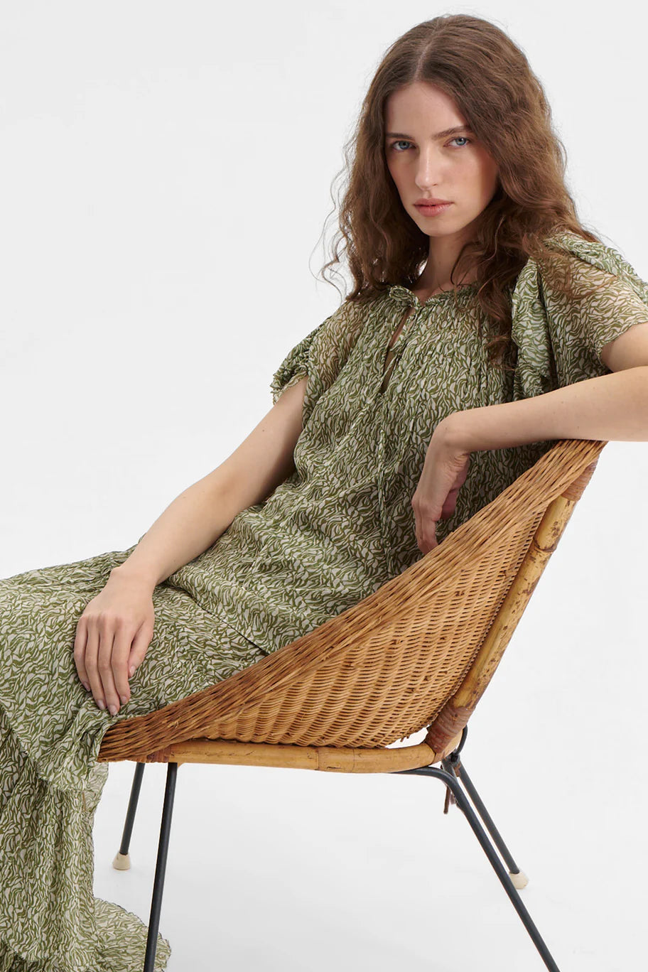 Woman in a green and white top and matching skirt  sitting on a wicker chair against a white background