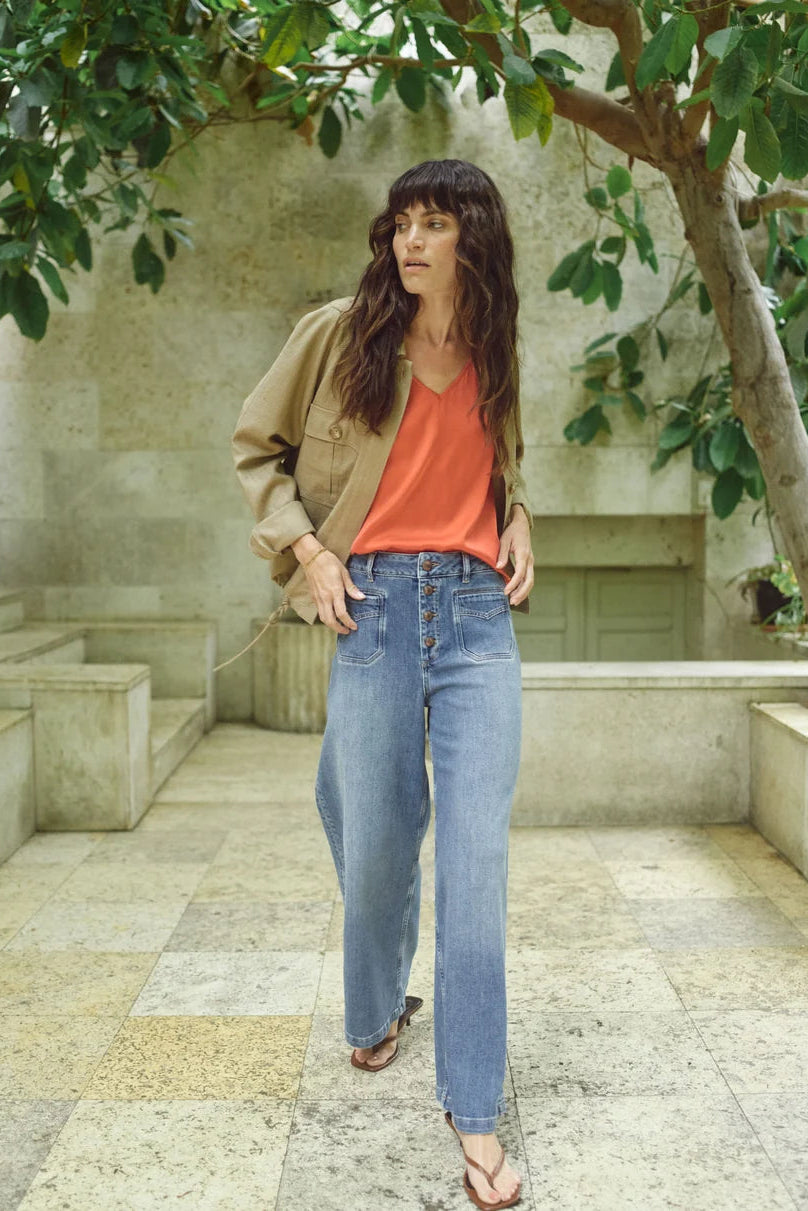 Woman standing outdoors in a garden setting wearing a beige jacket, orange top, and blue jeans.