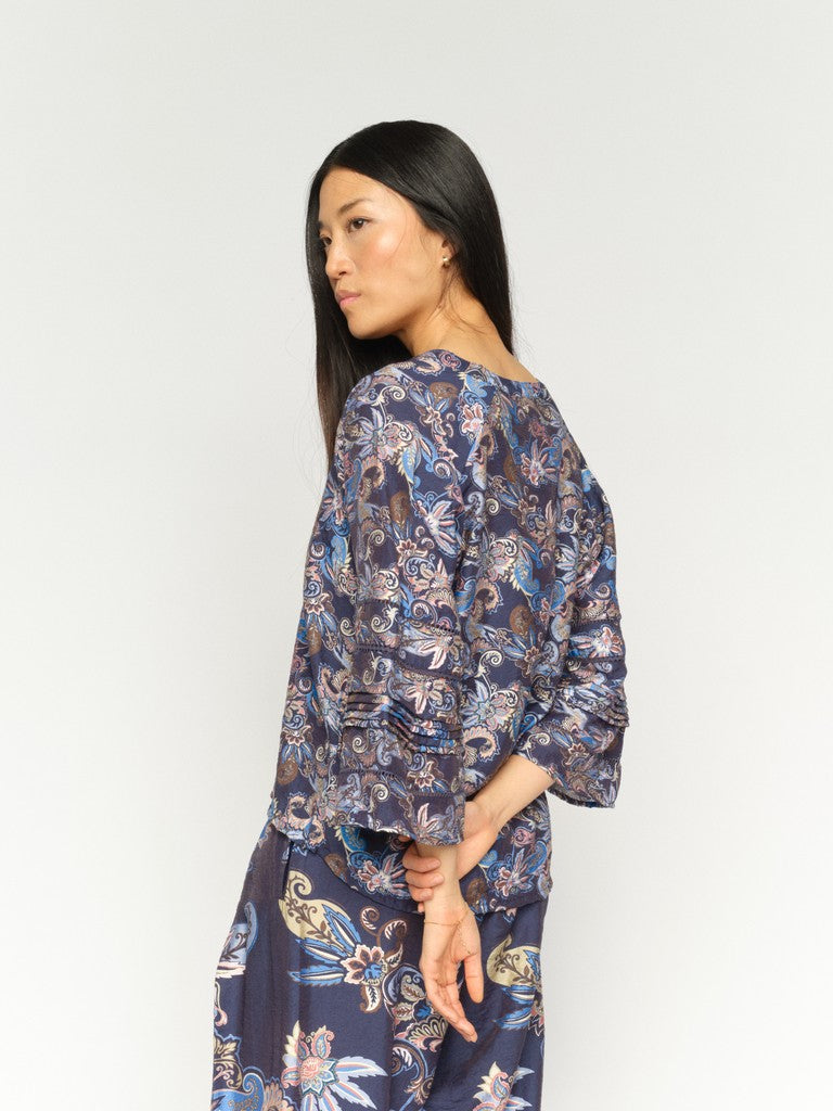 Woman shown from the side/back wearing a 3/4 sleeve v neck Paisley blue print blouse and matching pants on a white background. 
