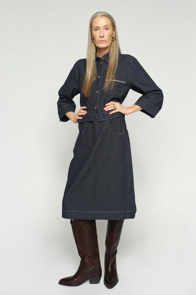 Model wearing a dark navy blue denim skirt with a fine white micro-dot pattern, featuring front patch pockets and a button closure with matching cropped jacket.  shown against a plain white background.