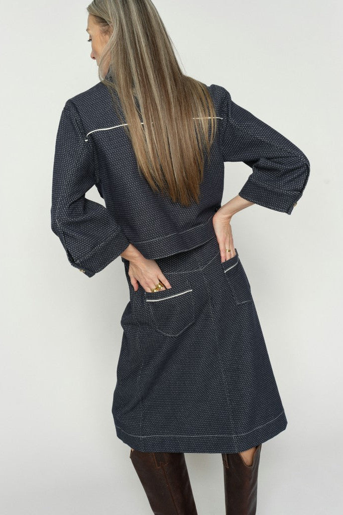 Model shown from the back wearing a dark navy blue denim skirt with a fine white micro-dot pattern, featuring front patch pockets and a button closure at the waistband, shown against a plain white background.