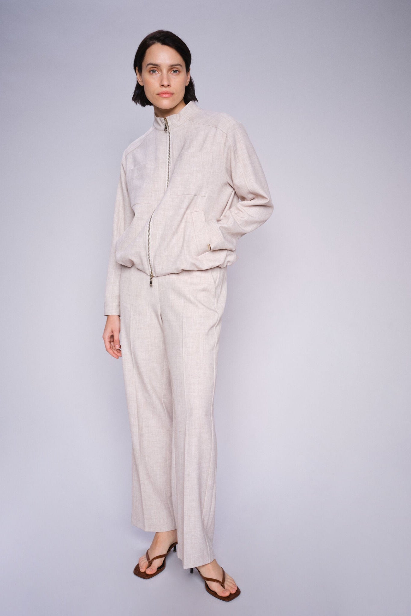 Model wearing beige straight leg pant with front pleats and elasticated waist with matching jacket and sandals on a white background. 