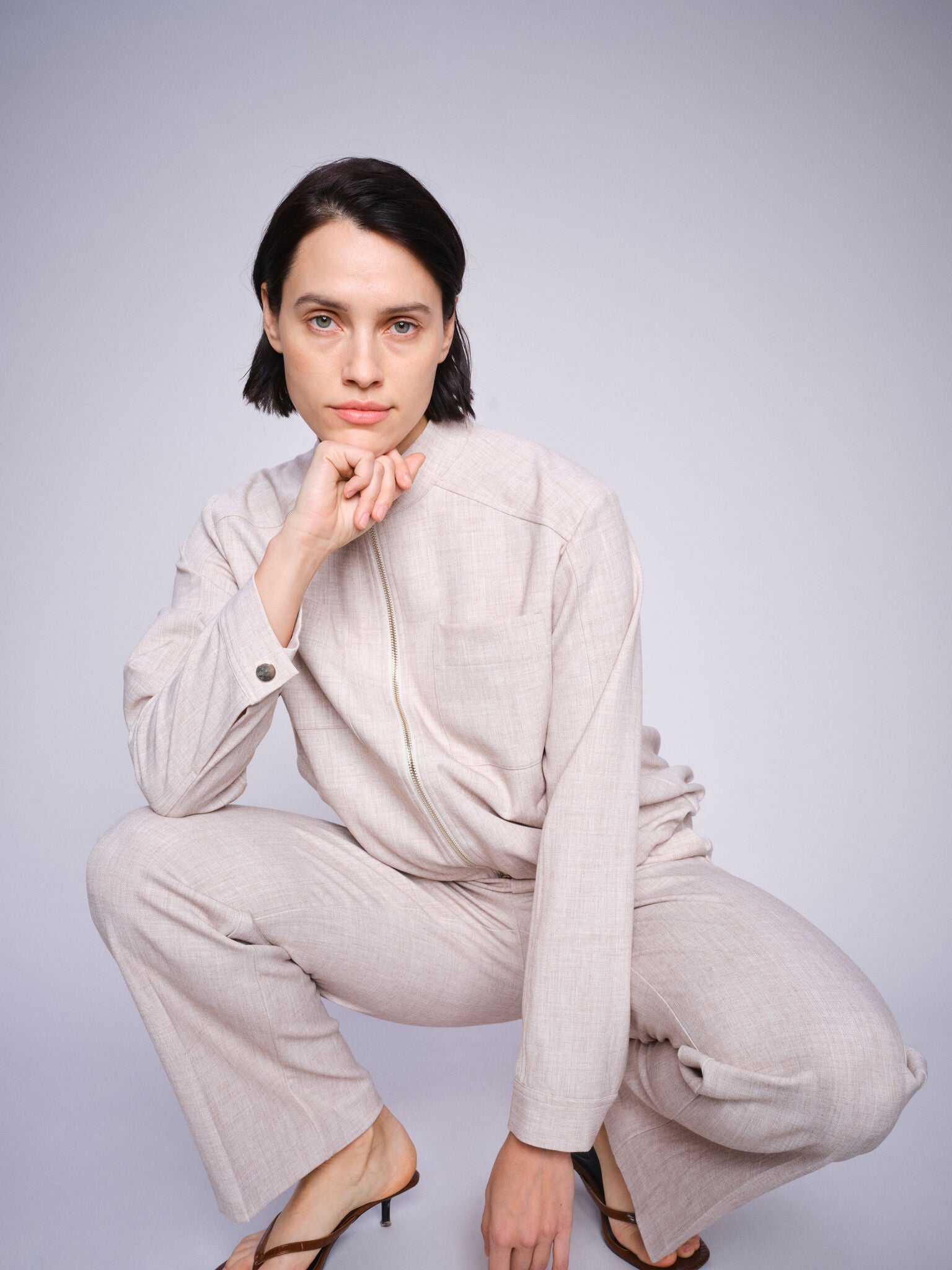 Model wearing beige straight leg pant with front pleats and elasticated waist with matching jacket paired with sandals on a white background. 