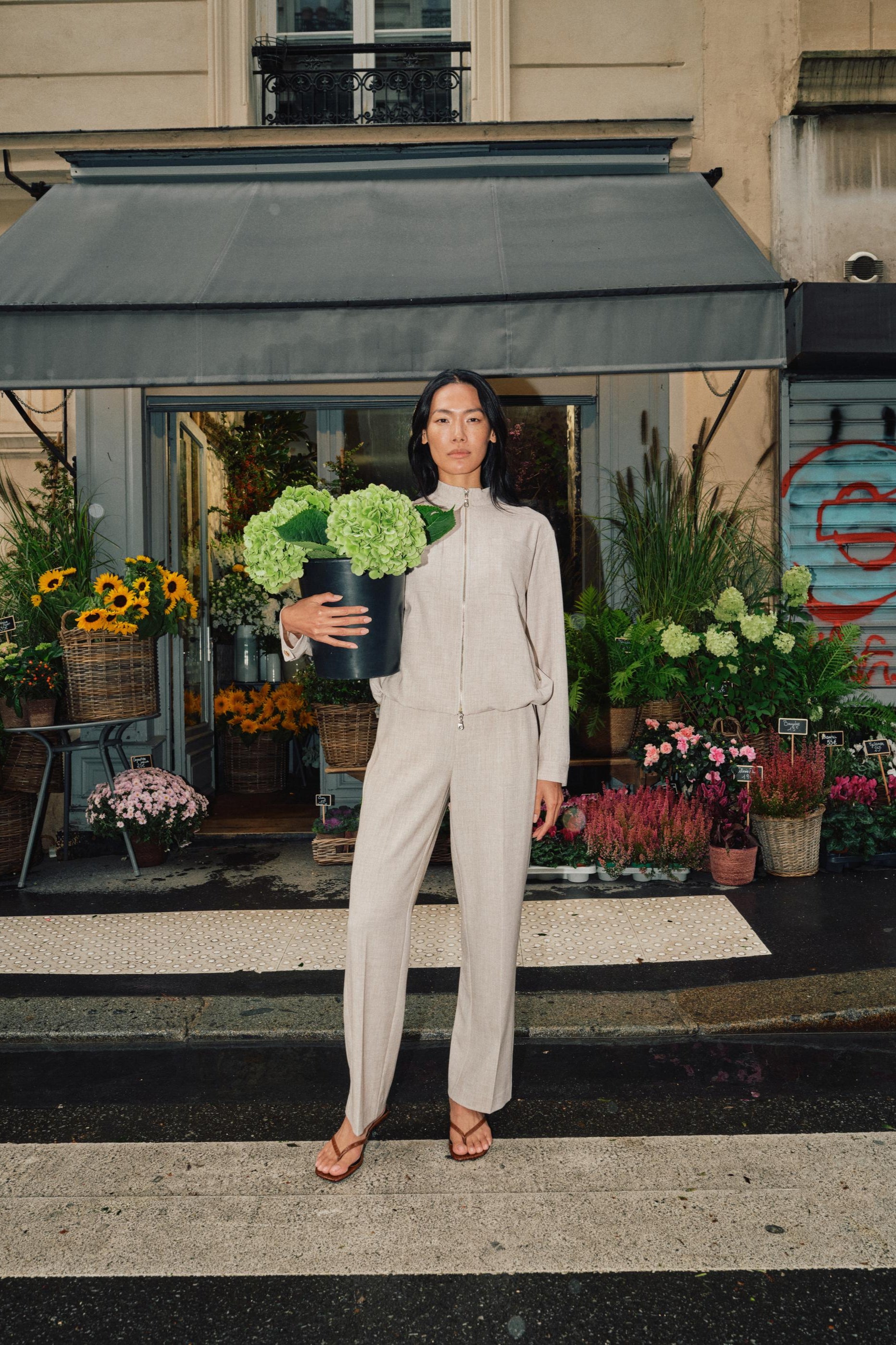 Model wearing beige straight leg pant with front pleats and elasticated waist at a flower market. 