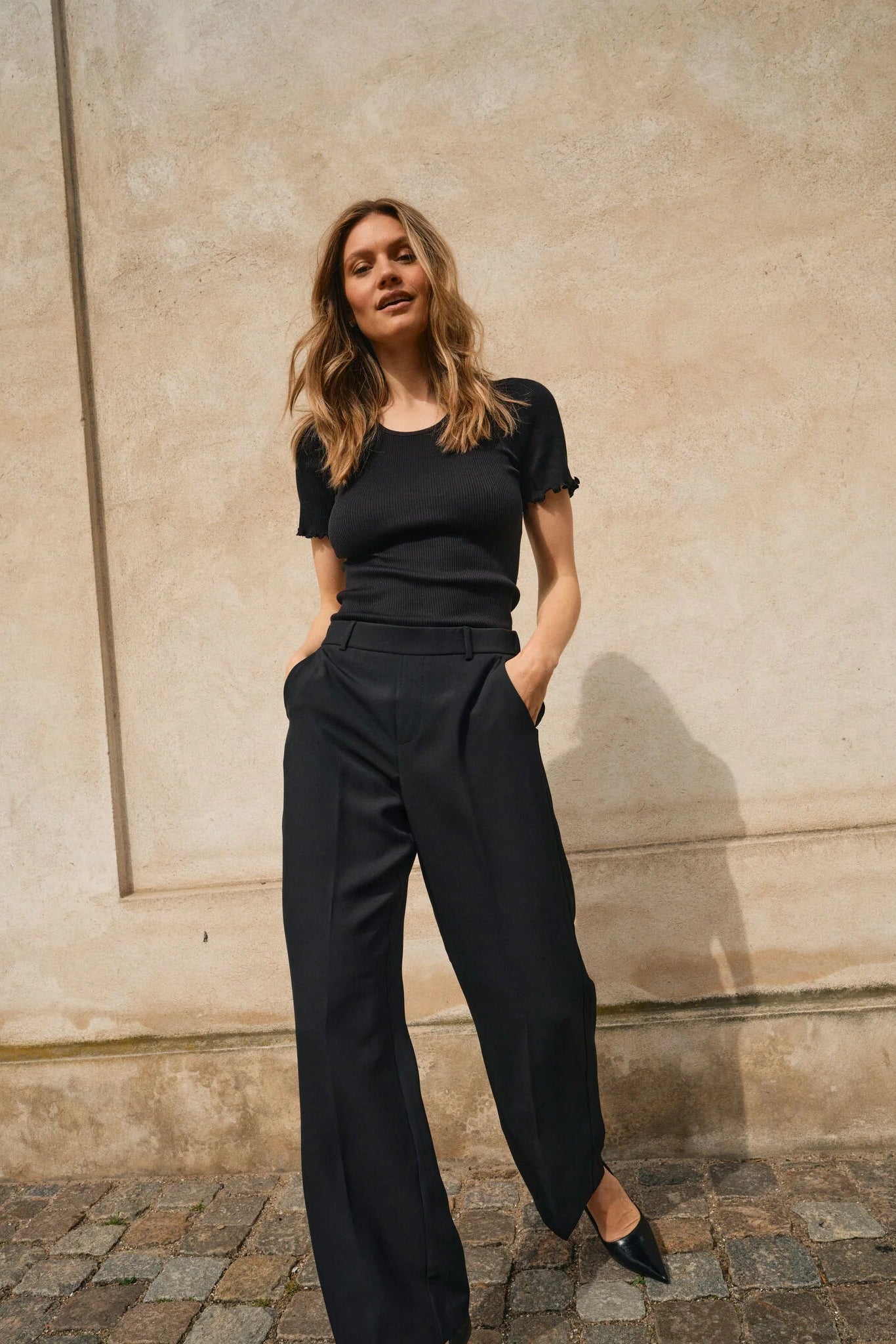 Woman wearing a black pants and black t shirt standing against a beige wall.