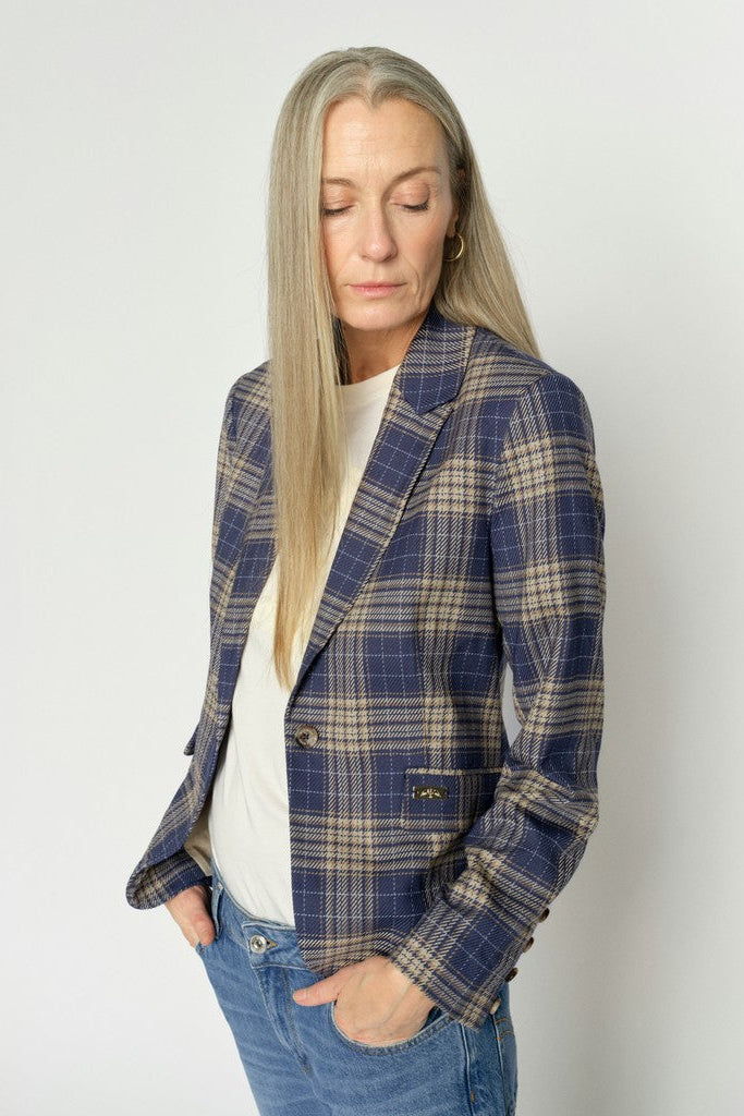A woman with long blonde hair wearing a navy blue and beige plaid blazer over a white top and blue jeans, standing against a light gray studio background.
