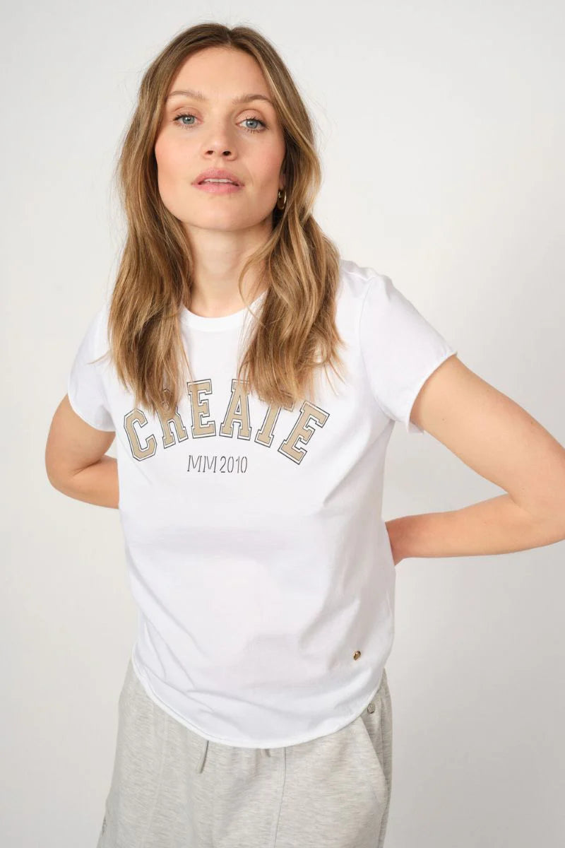 Woman wearing a white t-shirt with 'CREATE MM 2019' text in beige with light coloured pants on a plain background