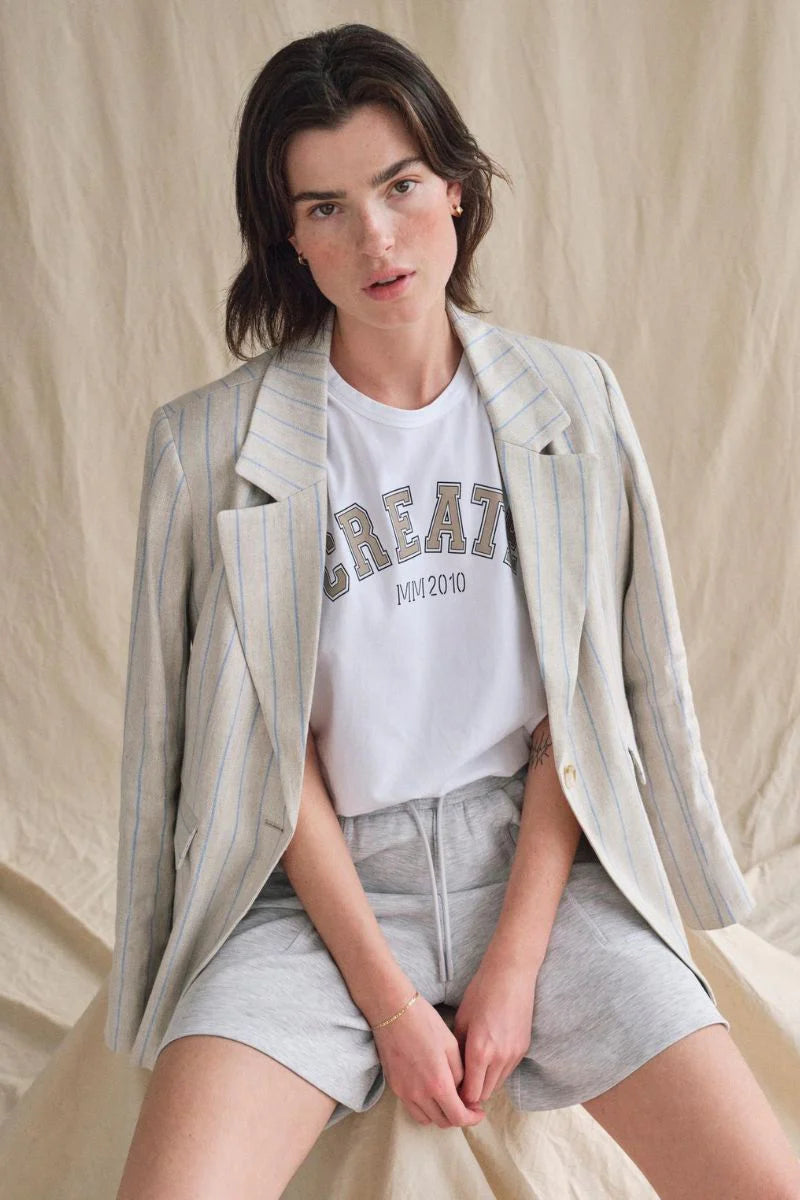 Model wearing white tee with "create" in beige across front centre.  Linen stripe jacket over shoulders and linen shorts on tan background. 
