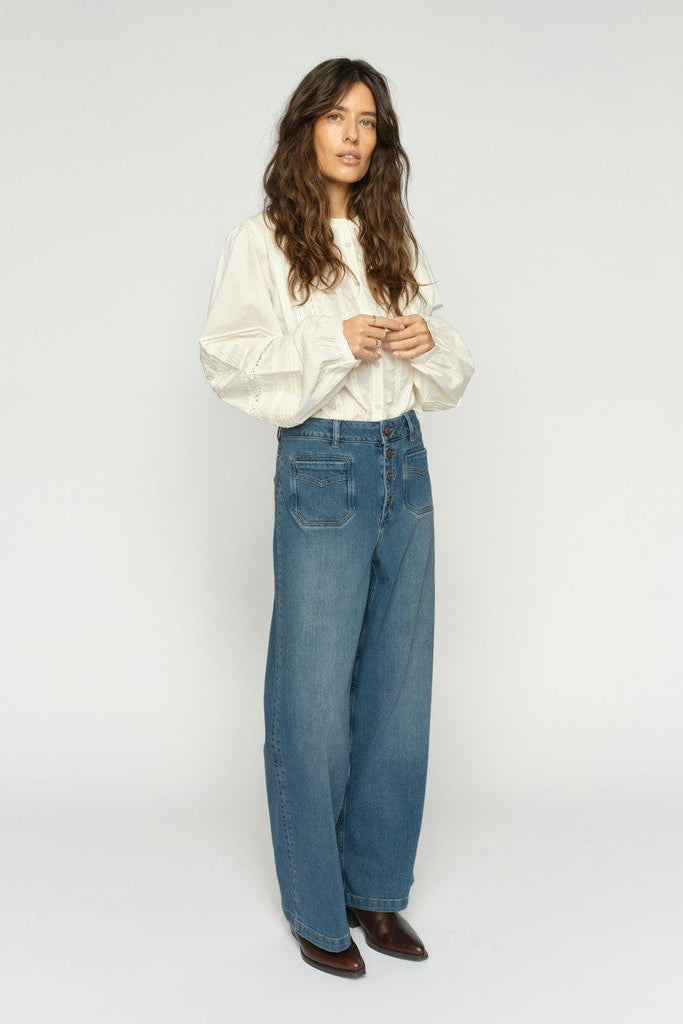 Women wearing a white long-sleeve blouse with wide leg blue denim jeans with exposed buttons and brown boots on a plain background.