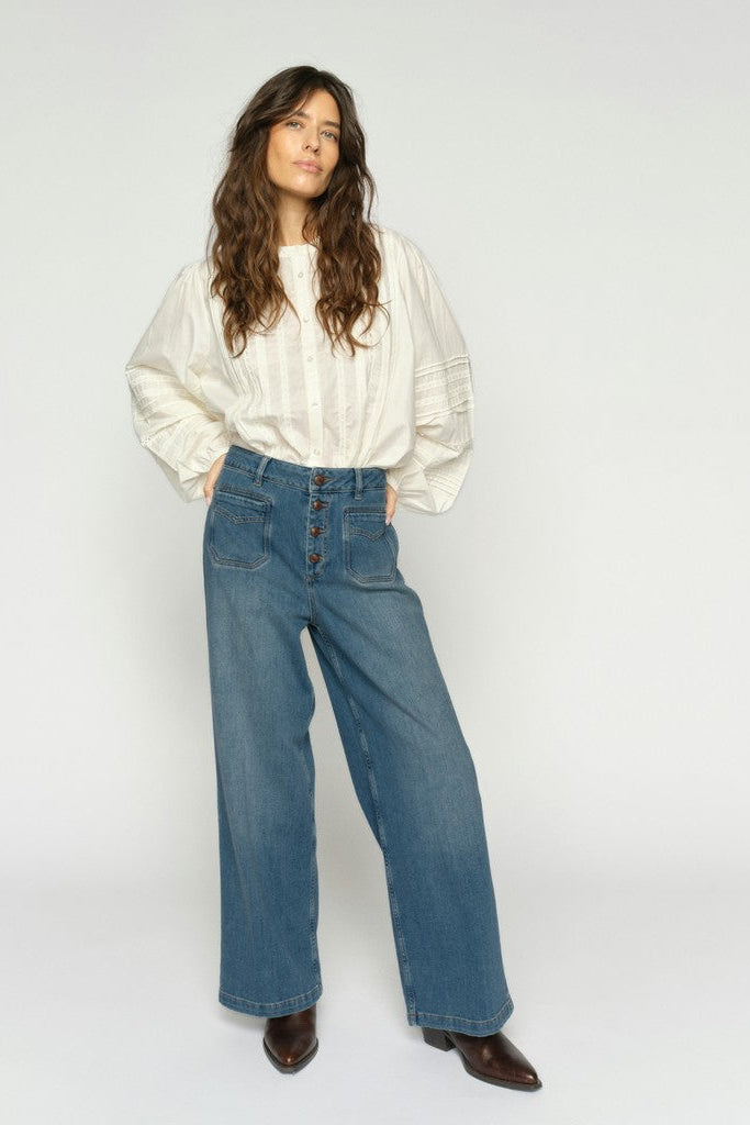 Women wearing a white long-sleeve blouse with wide leg blue denim jeans with exposed buttons and brown boots on a plain background.