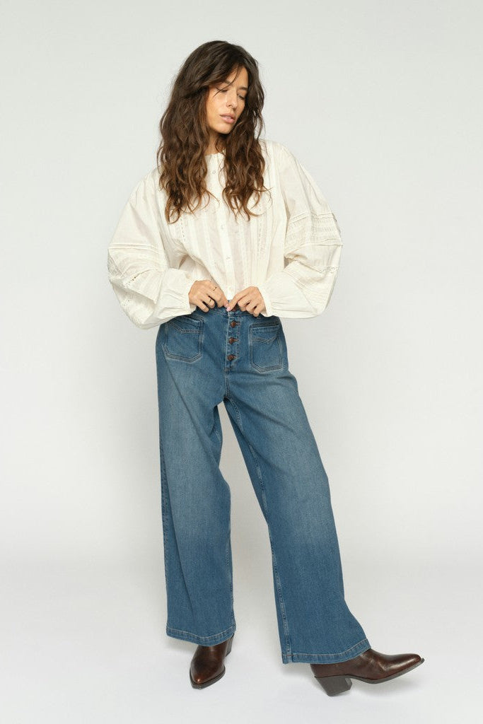 Women wearing a white long-sleeve blouse with wide leg blue denim jeans with exposed buttons and brown boots on a plain background.