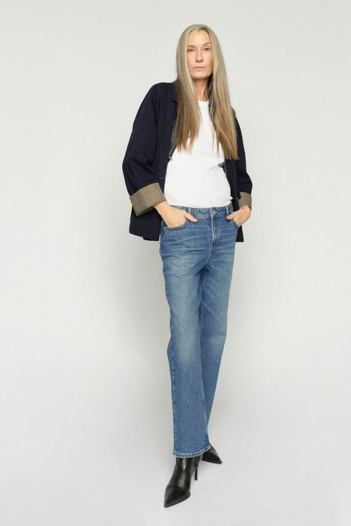 A woman is shown  wearing a Dark denim long-sleeve shirt with beige patterned cuffs, blue jeans, and black ankle boots, standing against a plain light background.