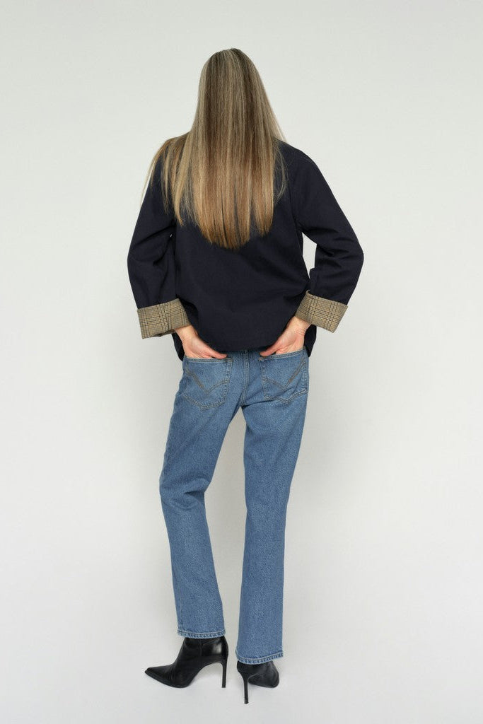 A woman is shown from behind wearing a Dark denim long-sleeve shirt with beige patterned cuffs, blue jeans, and black ankle boots, standing against a plain light background.