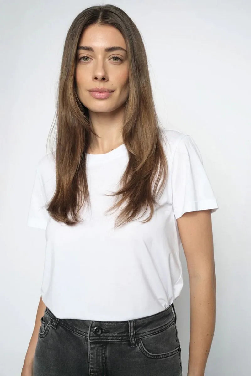 A woman with long brown hair wearing a plain white short-sleeve crewneck t-shirt and dark gray jeans, standing against a light gray studio background.