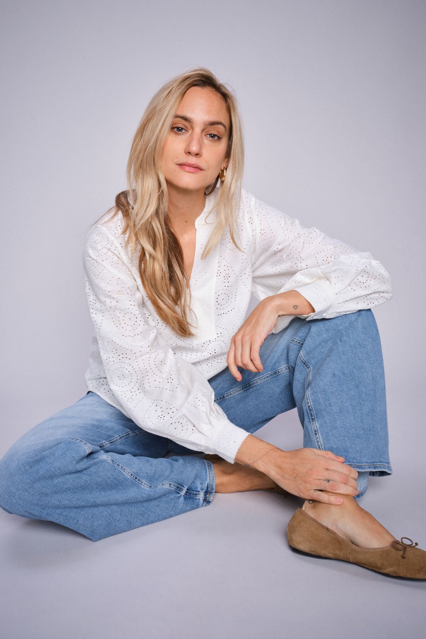 Woman wearing a white blouse and light blue jeans sitting down on a gray background