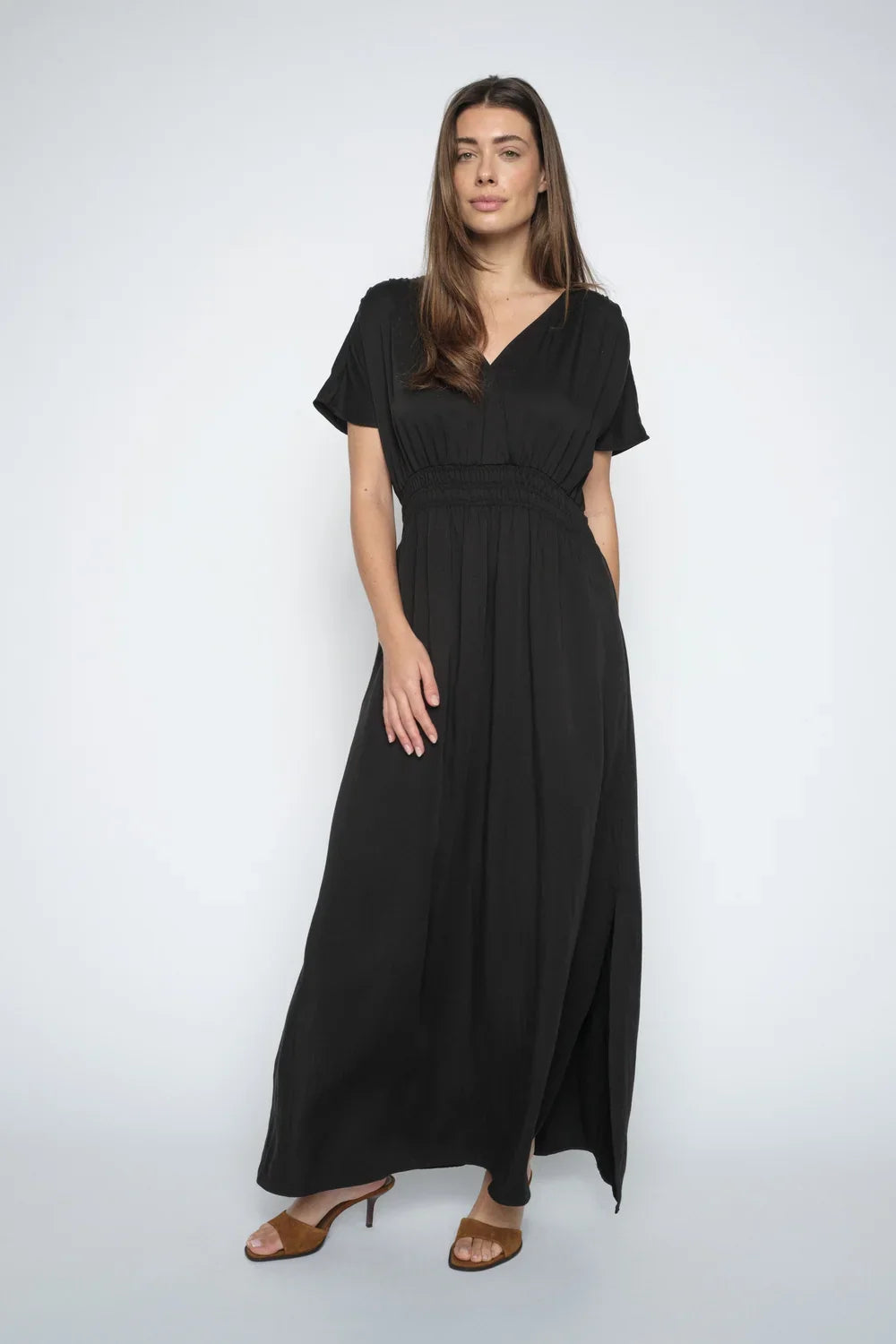A women wearing a black midi-length dress with short sleeves, a V-neckline, and a smocked elastic waist, with brown heels shown against a plain white background.