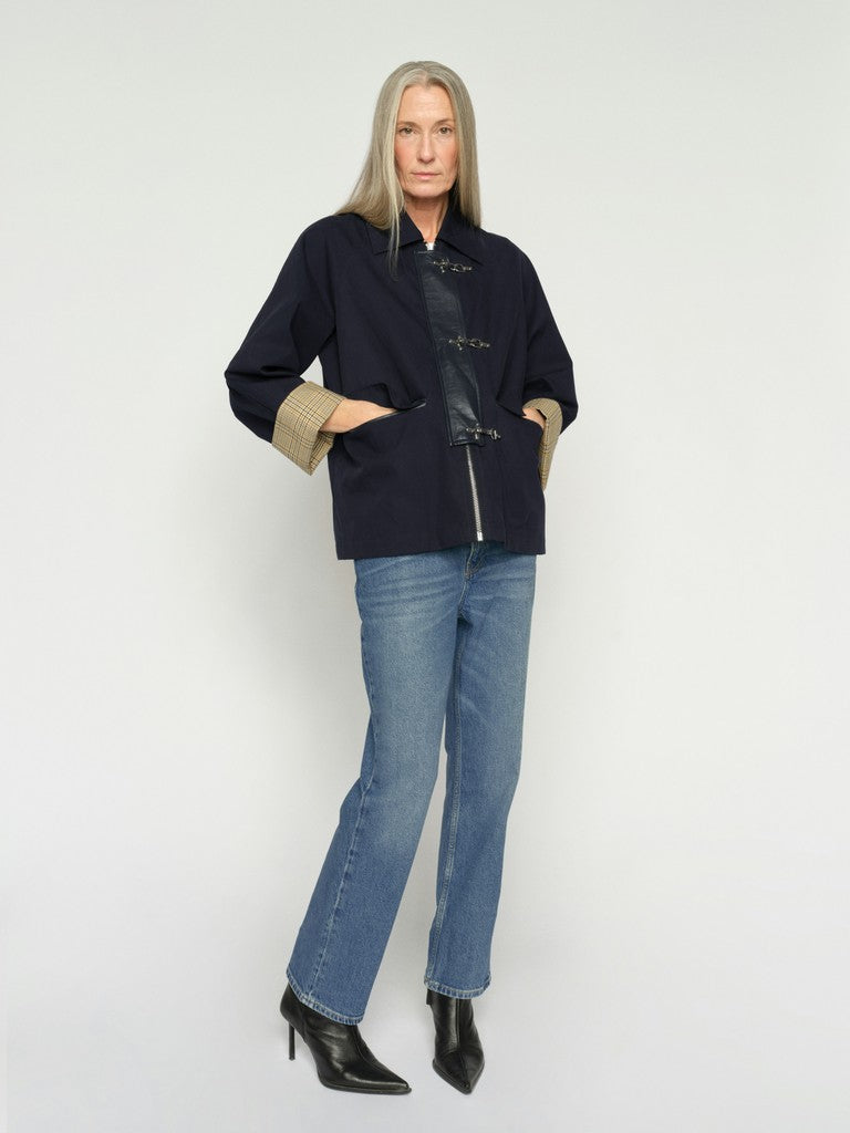 A woman with long blonde hair is shown wearing a dark navy blue jacket with a beige plaid cuff and blue jeans, against a plain light gray background.