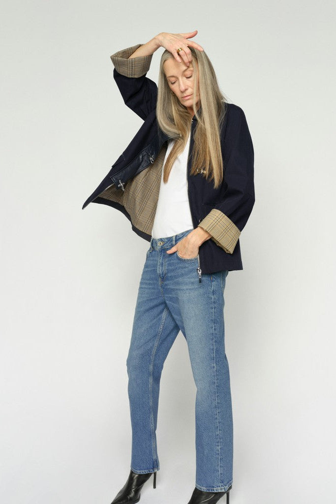 A woman with long blonde hair is shown wearing a dark navy blue jacket with a beige plaid cuff and blue jeans, against a plain light gray background.