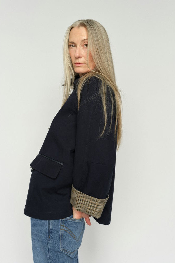 A woman with long blonde hair is shown from the side, wearing a dark navy blue jacket with a beige plaid cuff and blue jeans, against a plain light gray background.