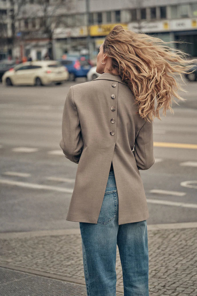 Person wearing a beige blazer with back button detailing and blue jeans walking on a city street.
