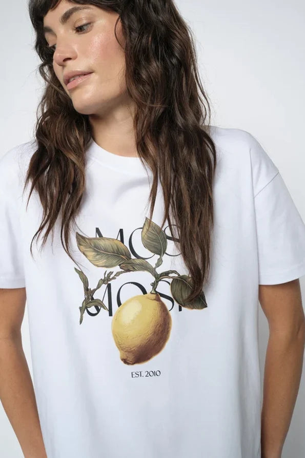 A woman wearing a white short-sleeve t-shirt with a graphic print of a lemon on a branch and the text 'Mosh Mosh est. 2010'.
