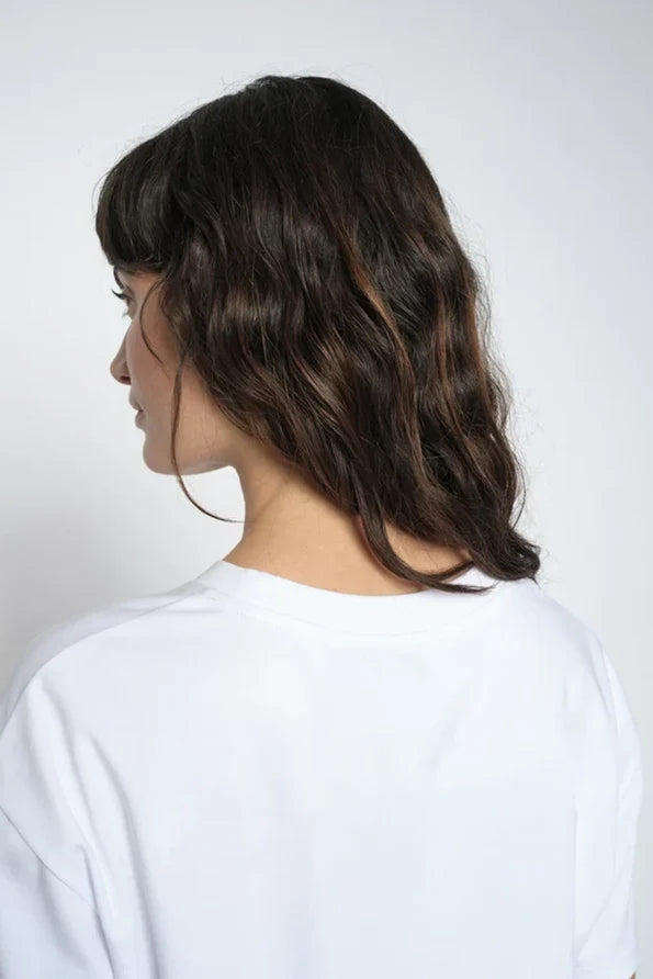 A woman shown from the back wearing a white crewneck t-shirt  with long brown hair set against a plain light grey background.