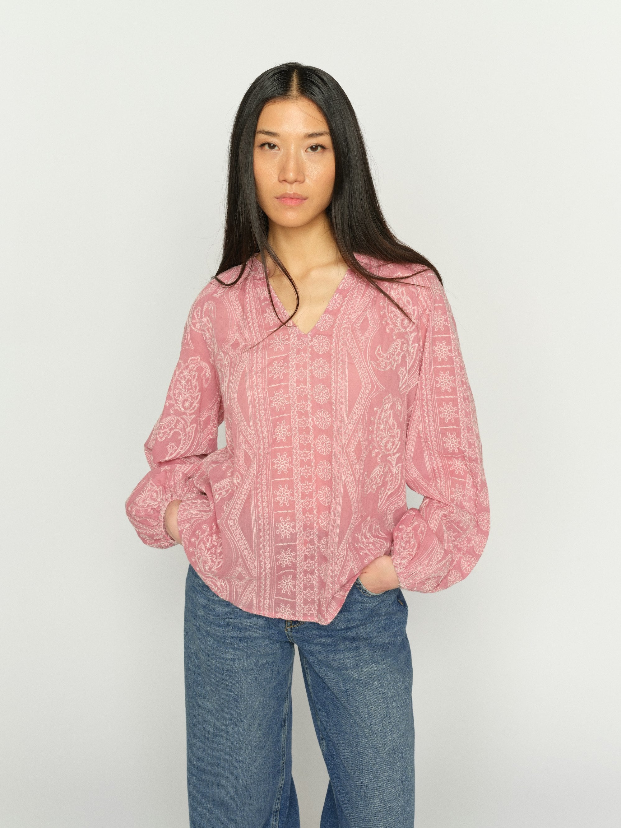 Women wearing a pink long-sleeve blouse with a white paisley and geometric pattern, featuring a split V-neckline and gathered cuffs, with blue denim jeans on a plain white background.