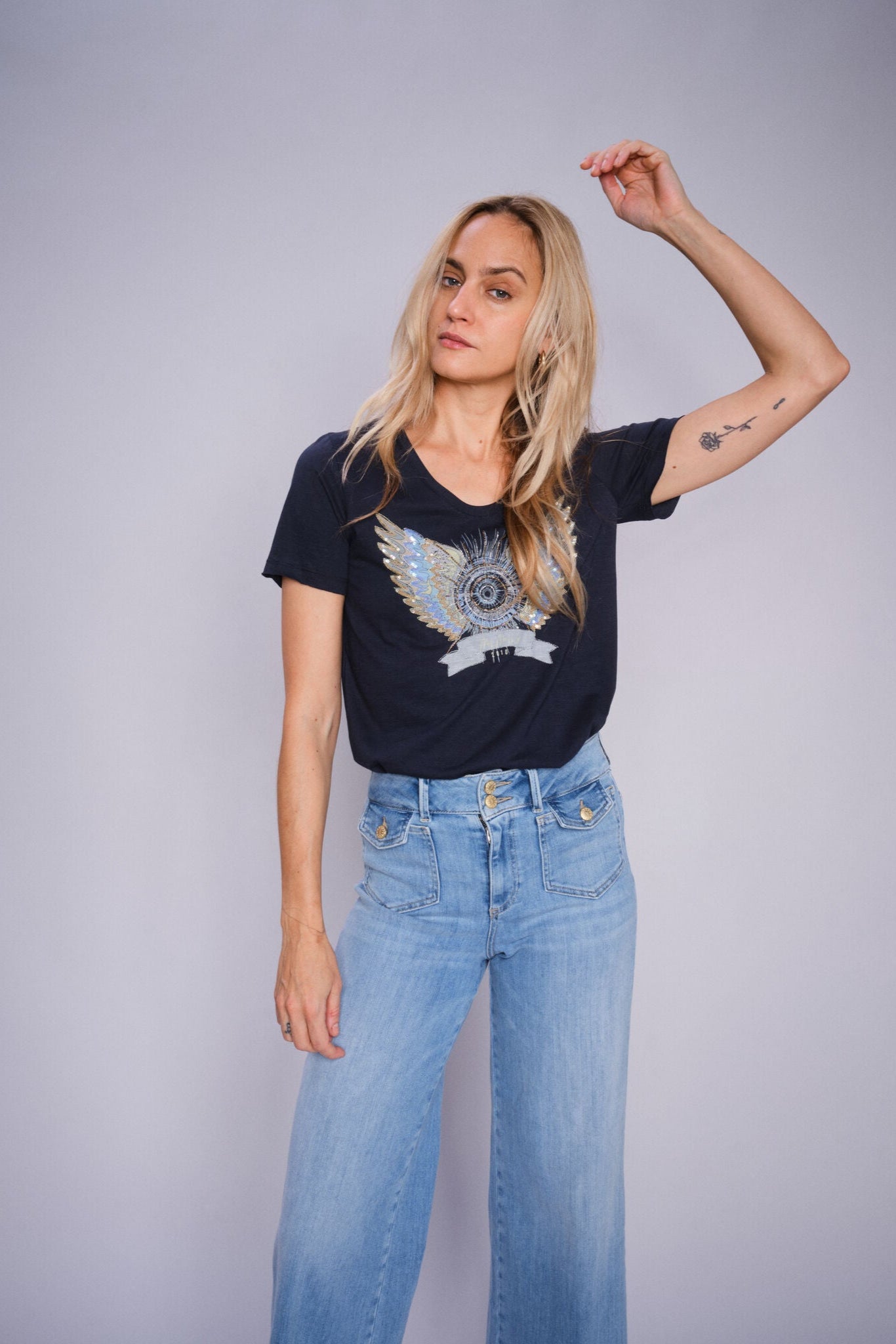 Model wearing navy crew neck t-shirt with a decorative angel wing design paired with light denim jeans. 