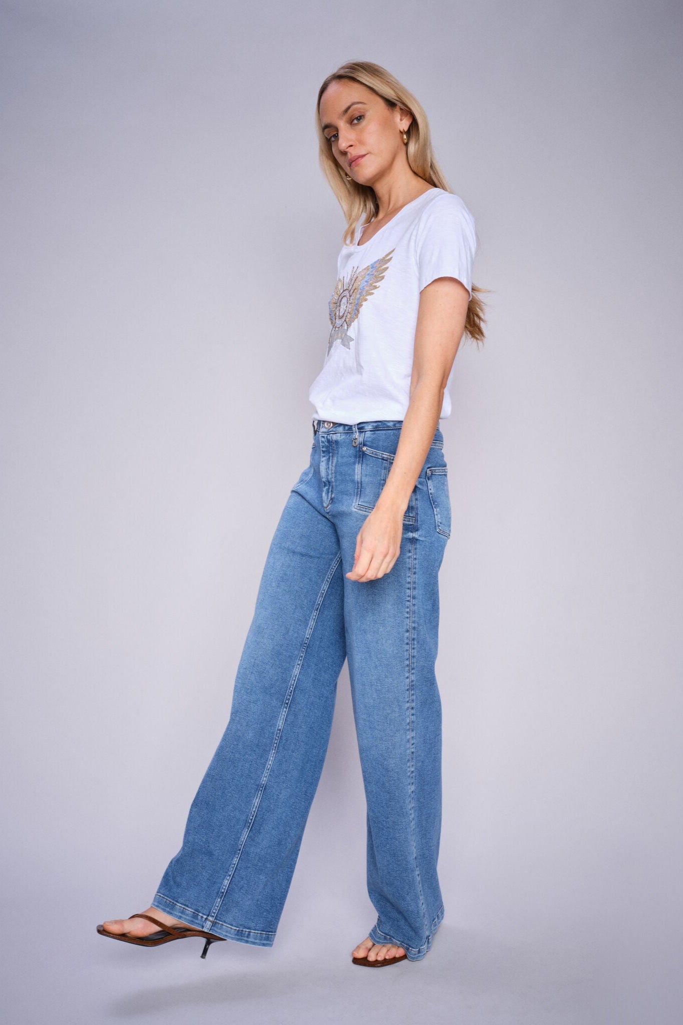 Model wearing white crew neck  t-shirt with gold and blue winged design and light denim jeans. 